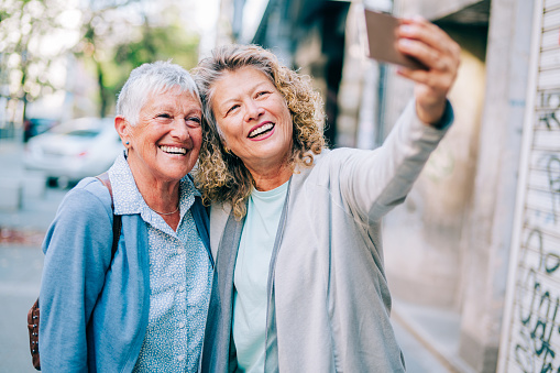 Shot of a two senior women using a phone while having a walk in the city. Two female mature friends making a selfie in foreign city.