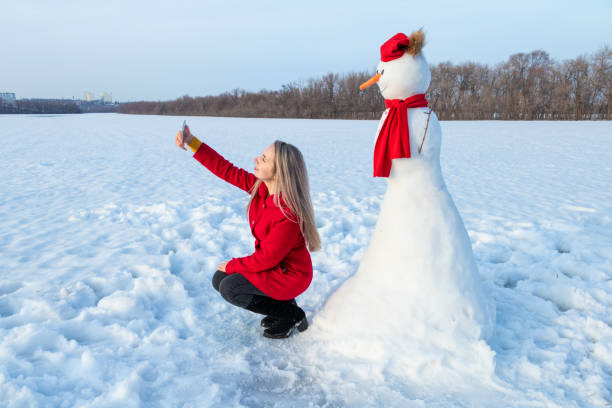 istock Attractive woman in red coat taking selfie with snowman using smartphone 1352531726