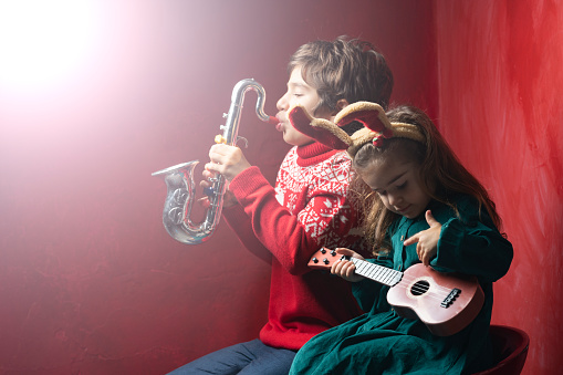Portrait of little brother and sister playing music for Christmas. The background is red colored wall. Boy is wearing a Christmas themed sweatshirt while girl is wearing a green dress. Shot with a full frame mirrorless camera.
