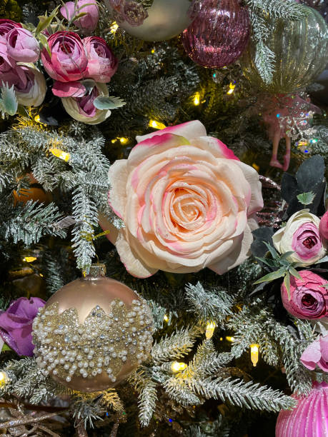 istock Image of individual artificial, rose flowerhead ornaments hanging on artificial Christmas tree branch, spruce needles covered with artificial spray snow, blurred white fairy lights background 1352538201