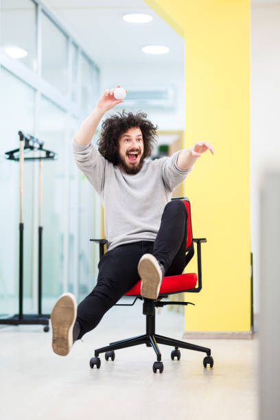 Young businessman with clumsy hair having fun at the office. Man on rolling chair throwing paper ball at colleagues Young businessman with clumsy hair having fun at the office. Man on rolling chair throwing paper ball at colleagues office chair stock pictures, royalty-free photos & images