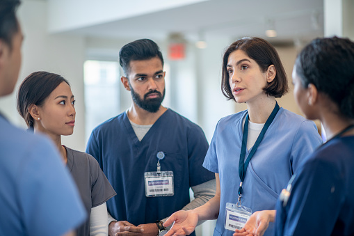 A small group of diverse medical professionals stand in the hallway for a brief meeting.  They are each wearing scrubs and focused on the conversation.
