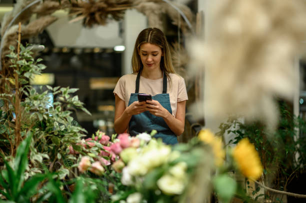 istock Florist using smartphone while surrounded with flowers and plants in a flower shop 1352554053