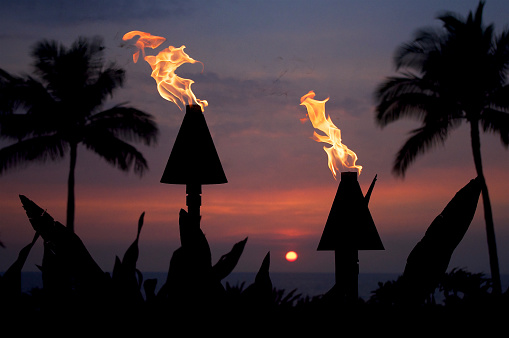 A colorful sunset silhouettes flaming tiki torches and palm trees on the Big Island of Hawaii
