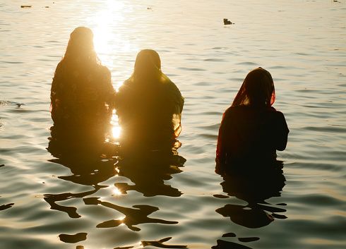 Kolkata, 11/10/2021: Silhouette of Hindu devotee women standing in knee deep water of Ganges, offering their prayer to Sun God 'Surya' at Ganges ghat during during Chhath Puja festival. The Chhath is ancient Hindu festival in Indian subcontinent, dedicated to praying the Sun God Surya.