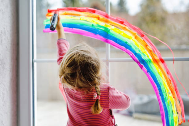 Adoralbe little toddler girl with rainbow painted with colorful window color during pandemic coronavirus quarantine. Child painting rainbows around the world with the words Let's all be well. stock photo