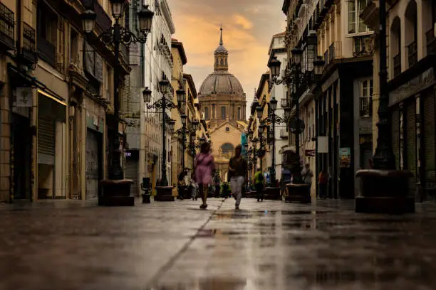 Low angle view of people walking along wet cobblestone pathway between stores and restaurants with Cathedral-Basilica of Our Lady of the Pillar in background.