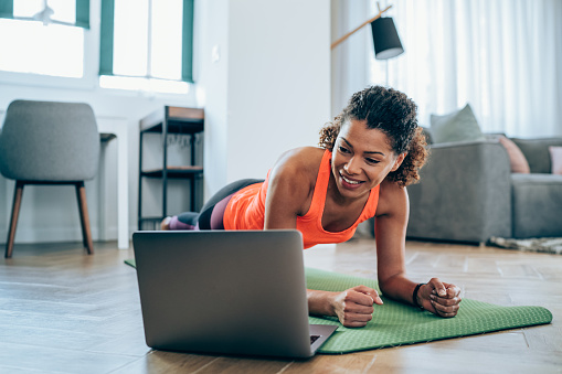 Woman doing exercises while watching workout video. Young woman using laptop while doing plank position. Young woman staying in front of her laptop and watching online sport lessons. Woman exercising and watching online fitness live streaming classes.