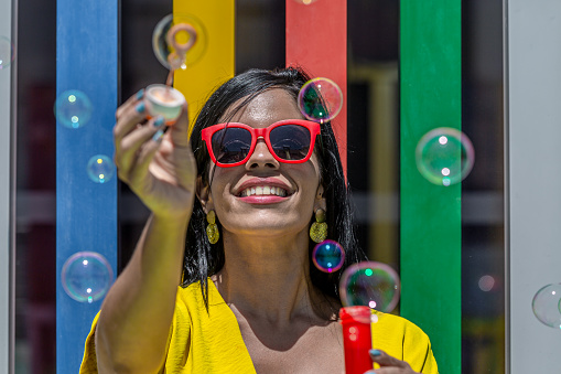 Front view of a Latina adult woman smiling and holding bubbles wand in a yellow dress and sunglasses against a background of colorful bars on a sunny day.