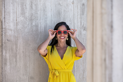 Front view near a column of a young Latina woman in yellow dress and sunglasses holding them with both hands and looking at camera against a wall at daytime