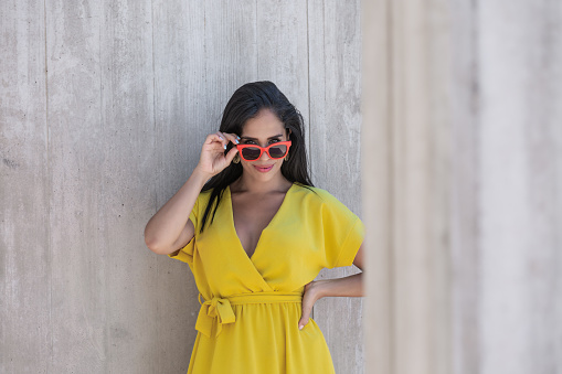 View near a column of a young Latina woman in yellow dress wearing sunglasses and holding them with one hand and looking at camera over the top of the glasses with the other hand on her waist.
