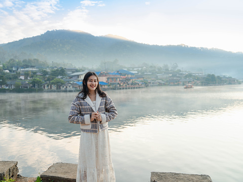 happy Asian woman in white dress and winter jacket standing at lakeside with fog rising on the lake view in the morning at Ban Rak Thai village, Mae Hong Son in Thailand.