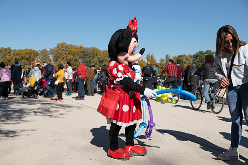 Madrid, Spain - Nov 09, 2021: A street performer dressed as Minie Mouse, gives balloons to beg in the Retiro park