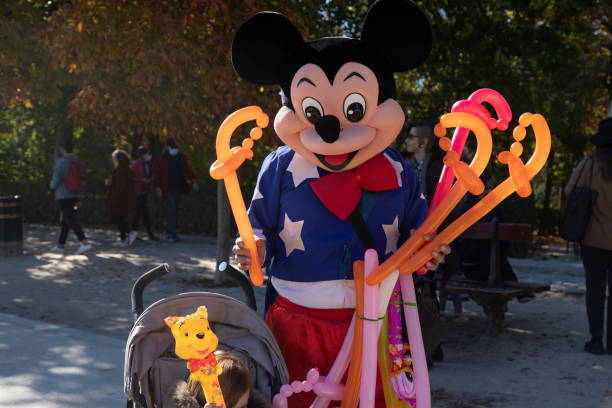 Madrid, Spain - Nov 09, 2021: A street performer dressed as Micky Mouse, poses with a child, making balloons to beg in the Retiro park