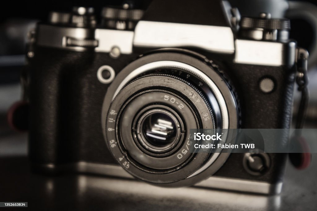 Detail of a vintage old 50mm USSR lens on a silver and black hybrid camera body on a grey desk top with coffee mug Studio pictures of an old vintage 50 mm USSR lens on a silver and black hybrid camera body on a grey desk top in a dark environment. Aluminum Stock Photo