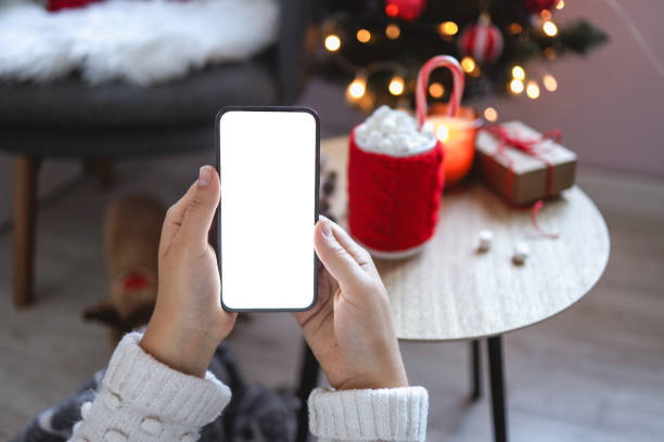 Close up smartphone with empty screen mockup on holiday background with Christmas tree, customer holding phone in hand, shopping online, purchasing gifts, searching information, browsing apps stock photo