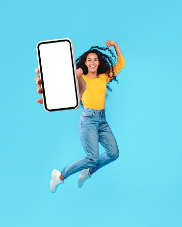 Happy Arab Female Jumping Up Showing Big Blank Smartphone Screen Advertising Great Application On Blue Background. Middle-Eastern Woman Holding Huge Cellphone For App Advertisement. Mockup, Collage
