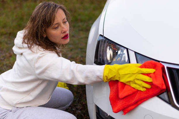 하얀 후드와 노란색 고무 장갑을 낀 예쁜 젊은 여성이 따뜻한 가을 날에 밝은 빨간 걸레가 달린 자동차의 헤드라이트를 닦아냅니다. 선택적 초점 - hood car headlight bumper 뉴스 사진 이미지