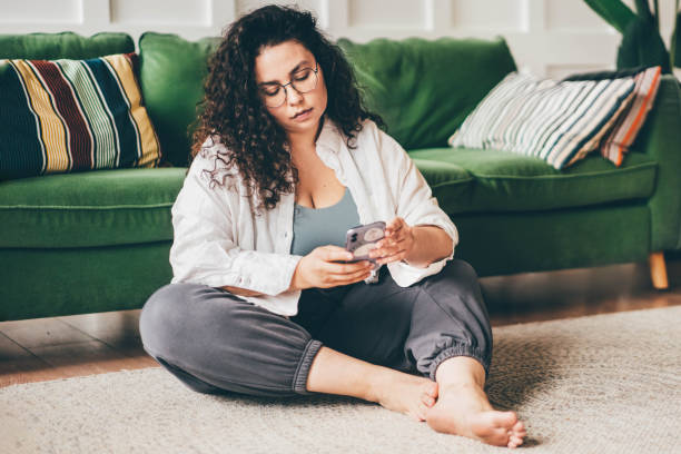 istock Curly haired plus size young woman wearing comfortable clothes in glasses clicks on smartphone screen sitting on floor mat in stylish room 1352655450