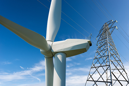 Closeup of a white wind turbine and high voltage tower (power line) against a clear blue sky with clouds and copy space. Renewable energy concept.