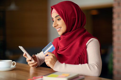 Young muslim lady in hijab using mobile phone and credit card while having coffee break at cafe, buying something online or paying for goods and services via mobile application, copy space