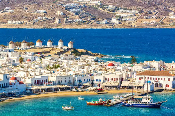 Photo of Chora port of Mykonos island with red church, famous windmills, ships and yachts during summer sunny day. Aegean sea, Greece