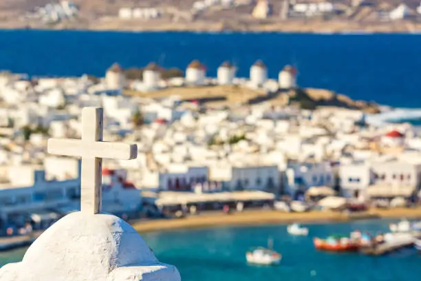 Photo of White Orthodox church cross against Chora port of Mykonos island with red church, famous windmills, ships and yachts during summer sunny day. Aegean sea, Greece