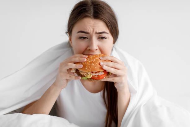 donna millenaria caucasica affamata affamata che mangia un grande hamburger seduta sul letto bianco in camera da letto - ingordigia foto e immagini stock