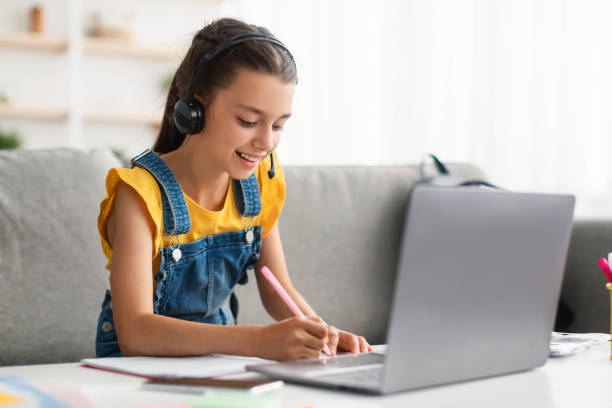 istock Positive schoolgirl sitting at desk, using laptop, writing in textbook 1352660542