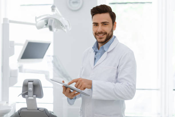 Attractive man dentist with digital tablet smiling at camera Attractive male dentist cheerful young bearded man with newest digital tablet smiling at camera, doctor using modern technologies while planning treatment, online appointment with dental clinic dentist stock pictures, royalty-free photos & images