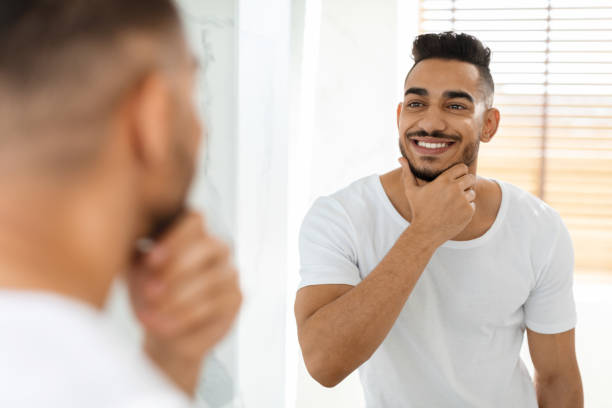 Male Facial Care. Handsome Arab Man Looking In Mirror And Touching Beard Male Facial Care. Handsome Arab Man Looking In Mirror In Bathroom And Touching Beard, Attractive Masculine Middle Eastern Guy Smiling To Reflection, Enjoying His Appearance, Selective Focus metrosexual stock pictures, royalty-free photos & images