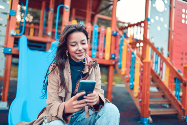 istock Smiling Female Sitting On Playground Slide And Using Smartphone 1352663620