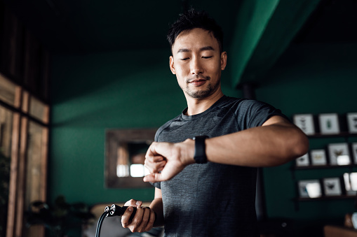Active young Asian man exercising at home, using fitness tracker app on smartwatch to monitor training progress and measuring pulse. Keeping fit and staying healthy. Health, fitness and technology concept