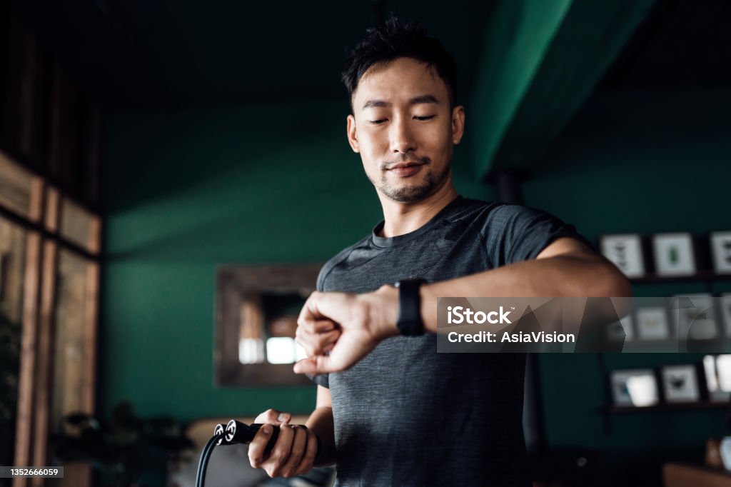 Active young Asian man exercising at home, using fitness tracker app on smartwatch to monitor training progress and measuring pulse. Keeping fit and staying healthy. Health, fitness and technology concept Exercising Stock Photo