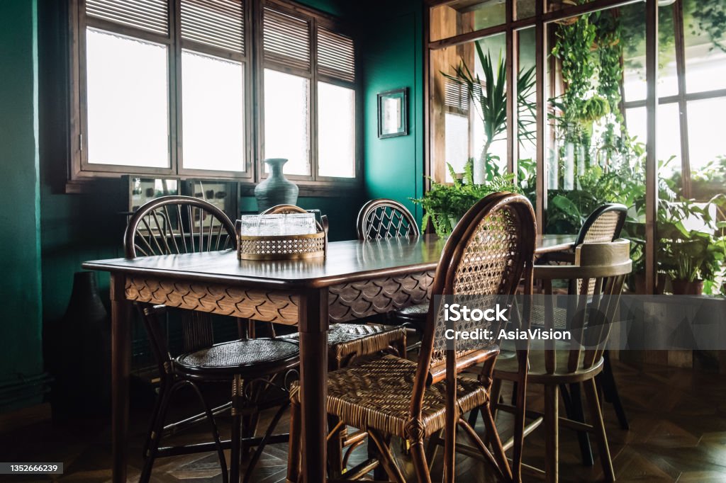A stylish dining room interior with brown coloured rattan furniture and wooden elements with dark green coloured wall. Decorated with plants Dining Room Stock Photo