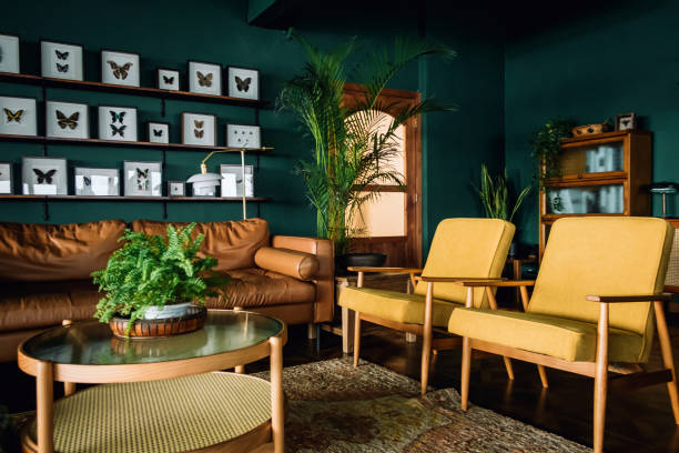 茶色と黄色の家具、ダークグリーンの壁が特徴の木製の要素を持つスタイリッシュなリビングルームのインテリア。植物や蝶の標本で飾られています - ボーホーシック ストックフォトと画像