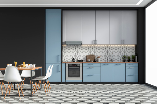 Empty modern kitchen with light blue and white high wooden kitchen cabinets with tiled pattern background,  ceiling led lights, dining table and  chairs. A  a high black plaster wall background on hexagon black and white tiled floor with copy space and windows on a side. Retro style of 70's. 3D rendered image.