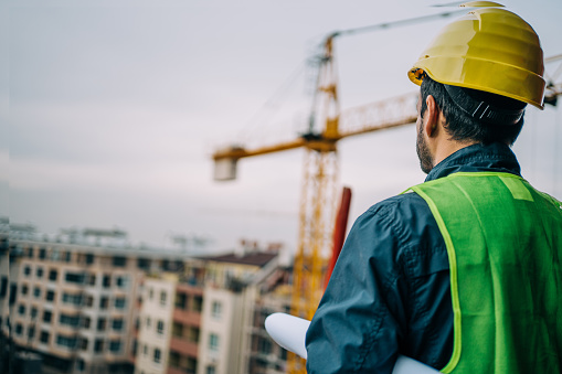 Shot of construction worker in uniform standing at construction site with crane in background. Construction engineer looking at new building while holding blueprint in hands.