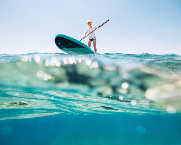Under the water view angle to the smiling blonde teenager, boy rowing stand up paddleboard. Active family summer vacation time near the sea concept image. Under the water view angle to the smiling blonde teenager, boy rowing stand up paddleboard. Active family summer vacation time near the sea concept image. paddleboard stock pictures, royalty-free photos & images