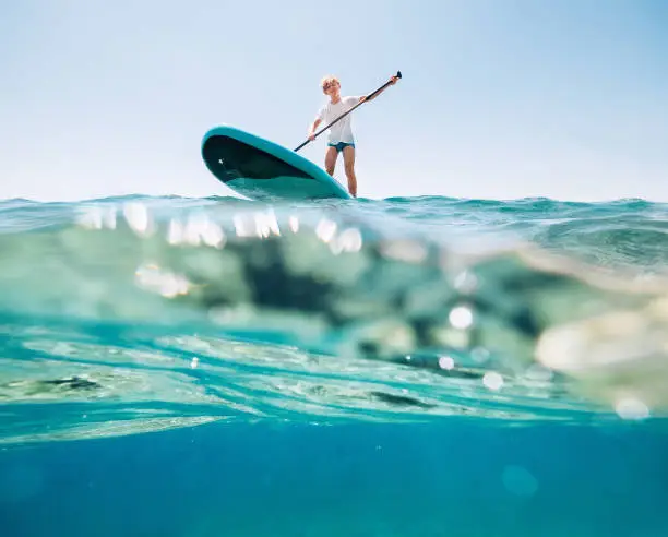 Photo of Under the water view angle to the smiling blonde teenager, boy rowing stand up paddleboard. Active family summer vacation time near the sea concept image.