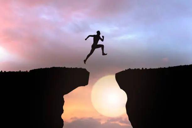 Photo of silhouette of businessman jumping over the cliff on sunset background, business concept idea