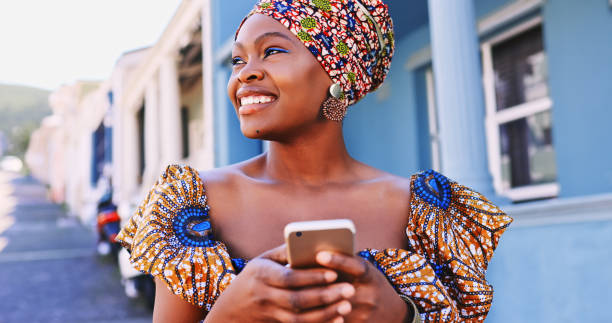 zdjęcie pięknej młodej kobiety w tradycyjnym afrykańskim stroju i używającej smartfona na miejskim tle - traditional clothing zdjęcia i obrazy z banku zdjęć