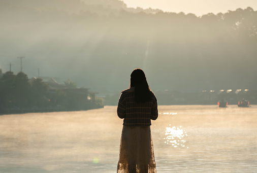 Detrás de una turista asiática de pie sola mira a la orilla del lago de Ban Rak Thai Village con niebla que se eleva en el lago por la mañana con rayos de sol y reflejo en Mae Hong Son, Tailandia photo