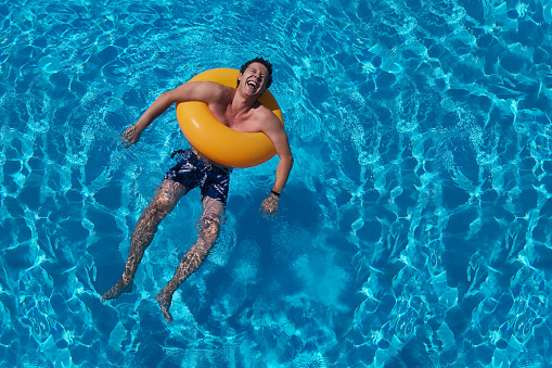 Orange circle and laughing Caucasian young man in summer pool. Saturated blue water background. Summer holidays, laughter, joy of life and cheerful man. Pool party