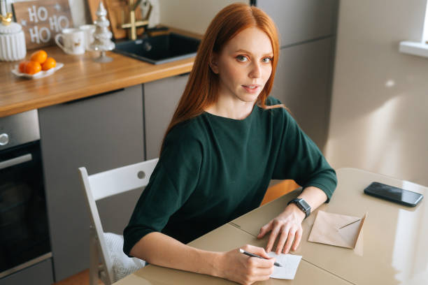 istock Attractive young woman writing handwritten letter sitting at table with envelope and mobile phone in room with modern light interior. 1352698018