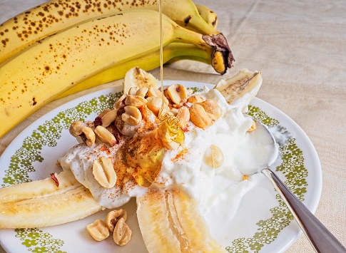 A banana split alternative, Greek yogurt banana split with peanuts and cinnamon. Served on a small plate with spoon and light colored tablecloth. A healthy snack packed with nutrition.