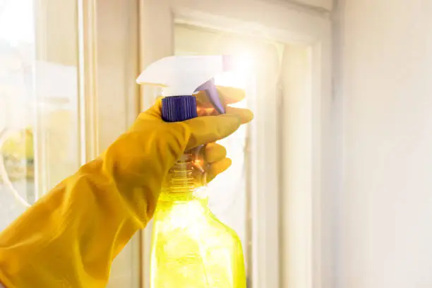 hand in yellow rubber gloves holds window cleaner by dirty glass. Selective focus. Blurred background.