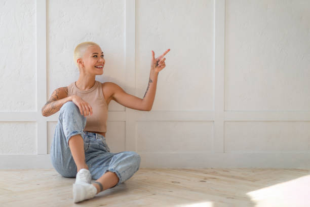 istock Young casual woman sitting on floor by white empty wall, pointing at copy space for advertisement, free space 1352711762