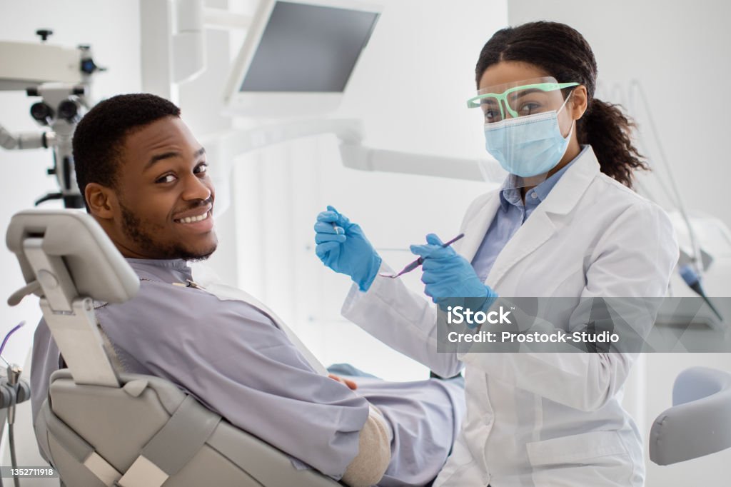 Professional Teeth Cleaning. Black Female Dentist Having Treatment With Male Patient Professional Teeth Cleaning. Black Female Dentist Having Treatment With Male Patient, Professional Stomatologist Woman In Medical Mask And Face Shield Holding Dental Tools And Looking At Camera Dentist Stock Photo
