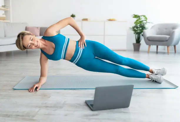 Photo of Smiling woman doing side plank on mat with pc